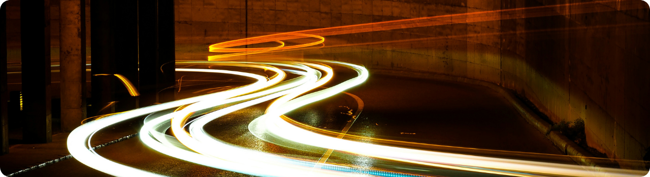 Long-exposure light trails curving through a concrete underpass at night, white and amber streaks on wet road.