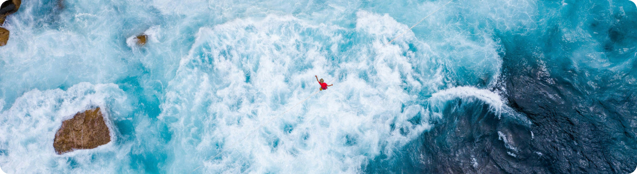 Aerial view of a person in a red top walking a slackline strung high above churning turquoise rapids.